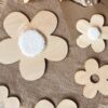Décoration murale composée de fleurs découpées en bois, disposées harmonieusement sur un mur clair. Les fleurs, de tailles variées, sont ornées de pompons blancs au centre, ajoutant une touche de douceur. À gauche et à droite, des plumes naturelles encadrent la composition, apportant une ambiance chaleureuse et naturelle. Créées et découpées en france.