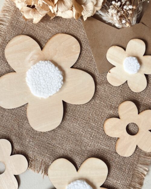 Décoration murale composée de fleurs découpées en bois, disposées harmonieusement sur un mur clair. Les fleurs, de tailles variées, sont ornées de pompons blancs au centre, ajoutant une touche de douceur. À gauche et à droite, des plumes naturelles encadrent la composition, apportant une ambiance chaleureuse et naturelle. Créées et découpées en france.